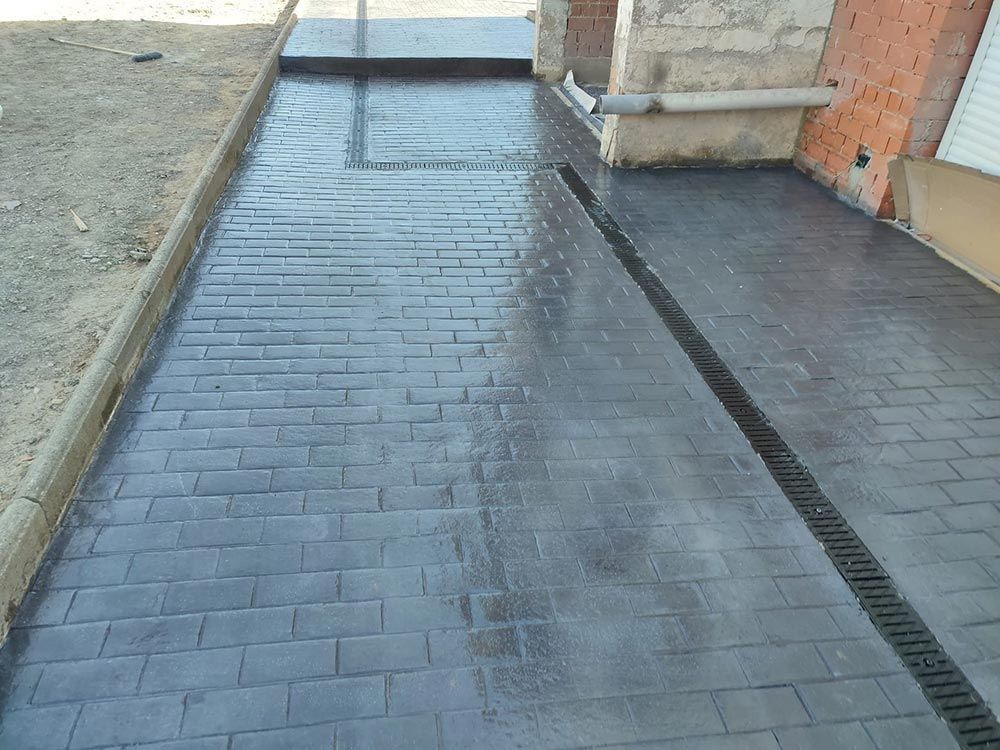 Entrada de vehículos de hormigón estampado gris con patrón de ladrillo y canal de drenaje.