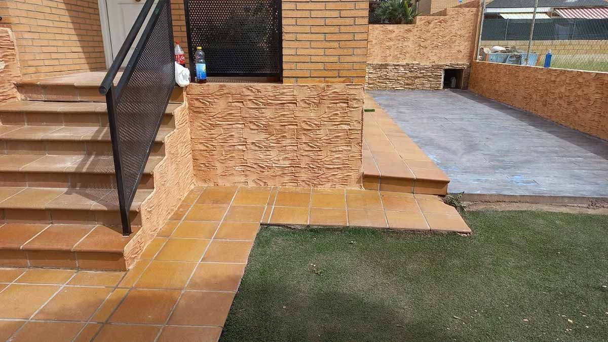 Exterior de casa de ladrillo con escaleras, pared texturizada, área pavimentada y césped.