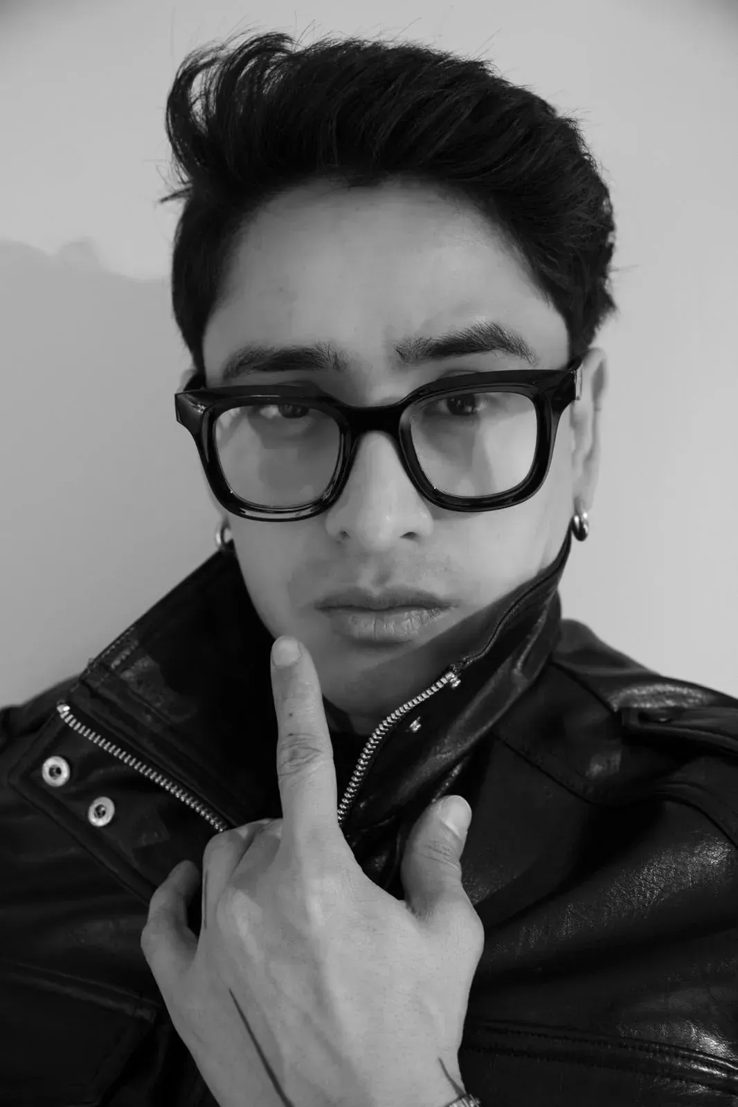 Persona con chaqueta negra y gafas, posando con un dedo en la barbilla frente a un fondo liso.