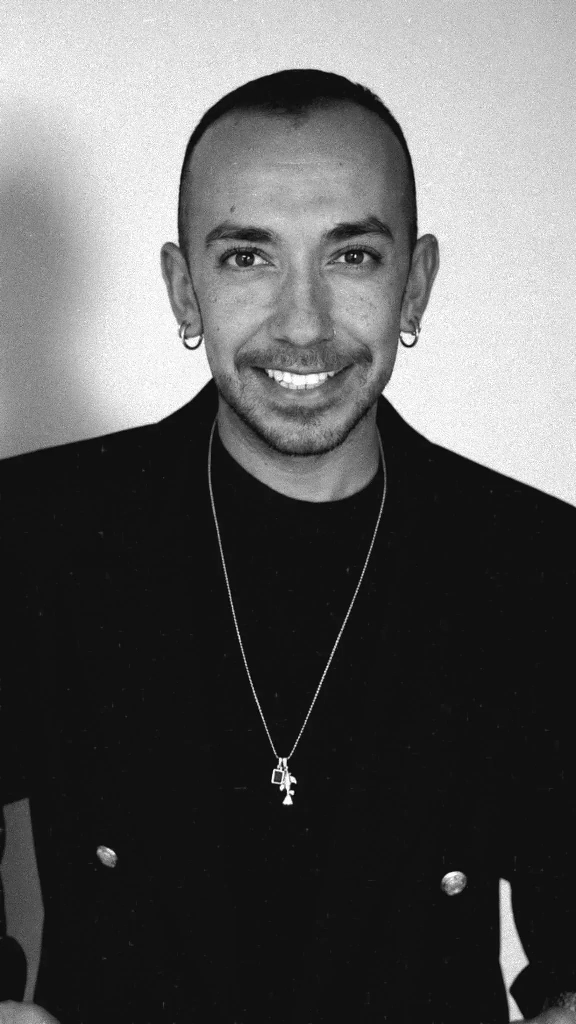 Persona sonriente con un jersey oscuro y un collar, de pie frente a un fondo liso.