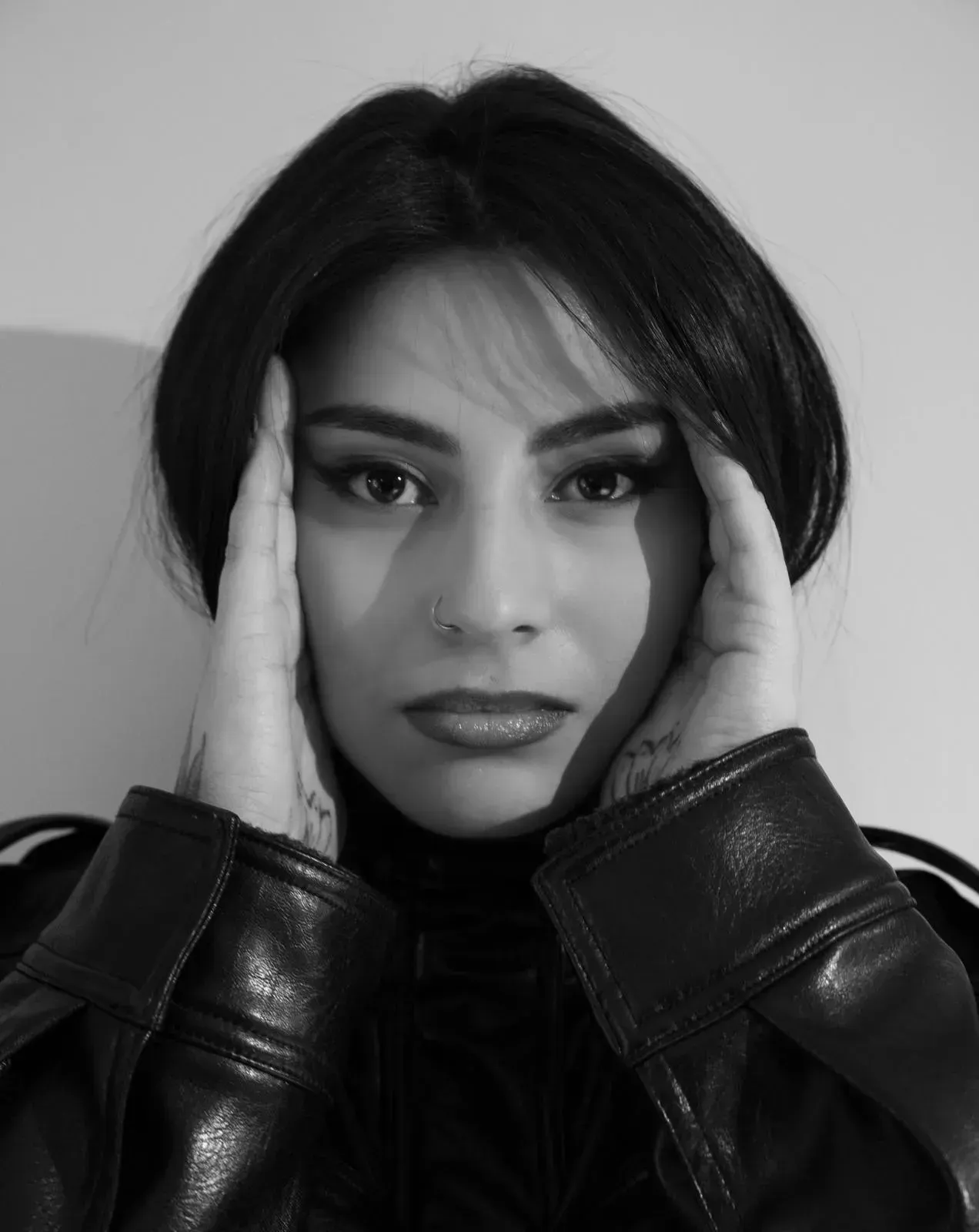 Retrato en blanco y negro de una persona con una chaqueta de cuero, con las manos enmarcando el rostro, mirando a la cámara.