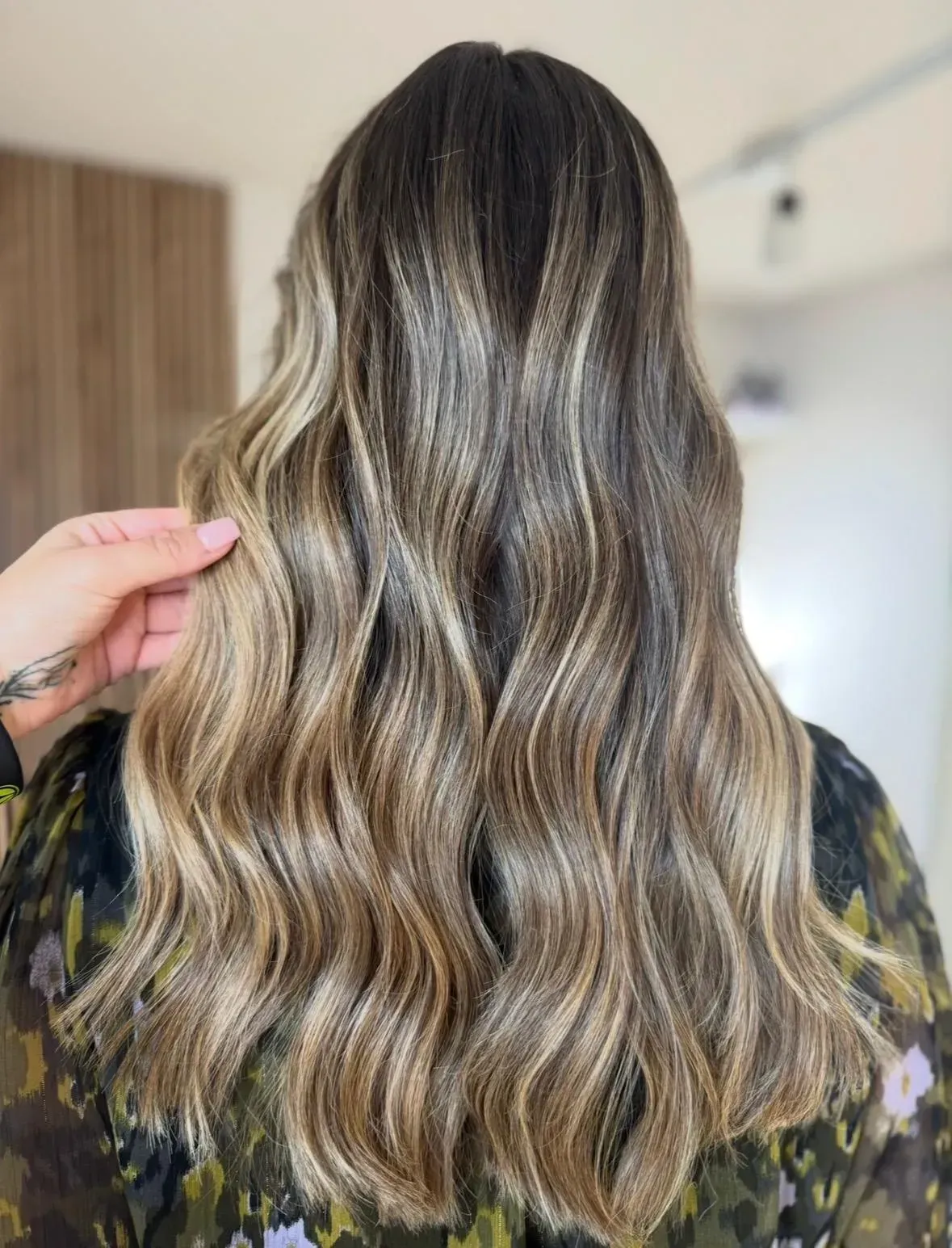 Cabello largo, castaño y ondulado, con mechas rubias, sostenido por una mano frente a un fondo interior desenfocado