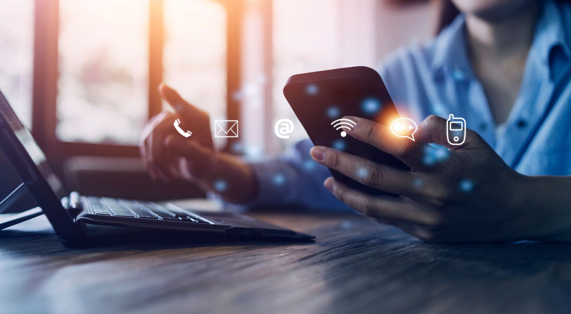 Persona que usa un teléfono inteligente y una computadora portátil en un escritorio, con íconos digitales superpuestos.