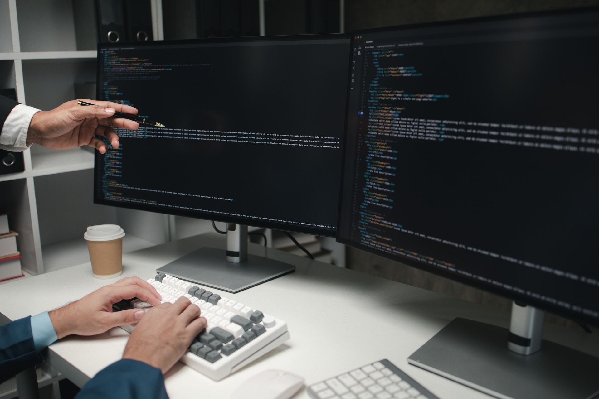Una persona señala un código en un monitor mientras otra escribe en un teclado, en un entorno de oficina.