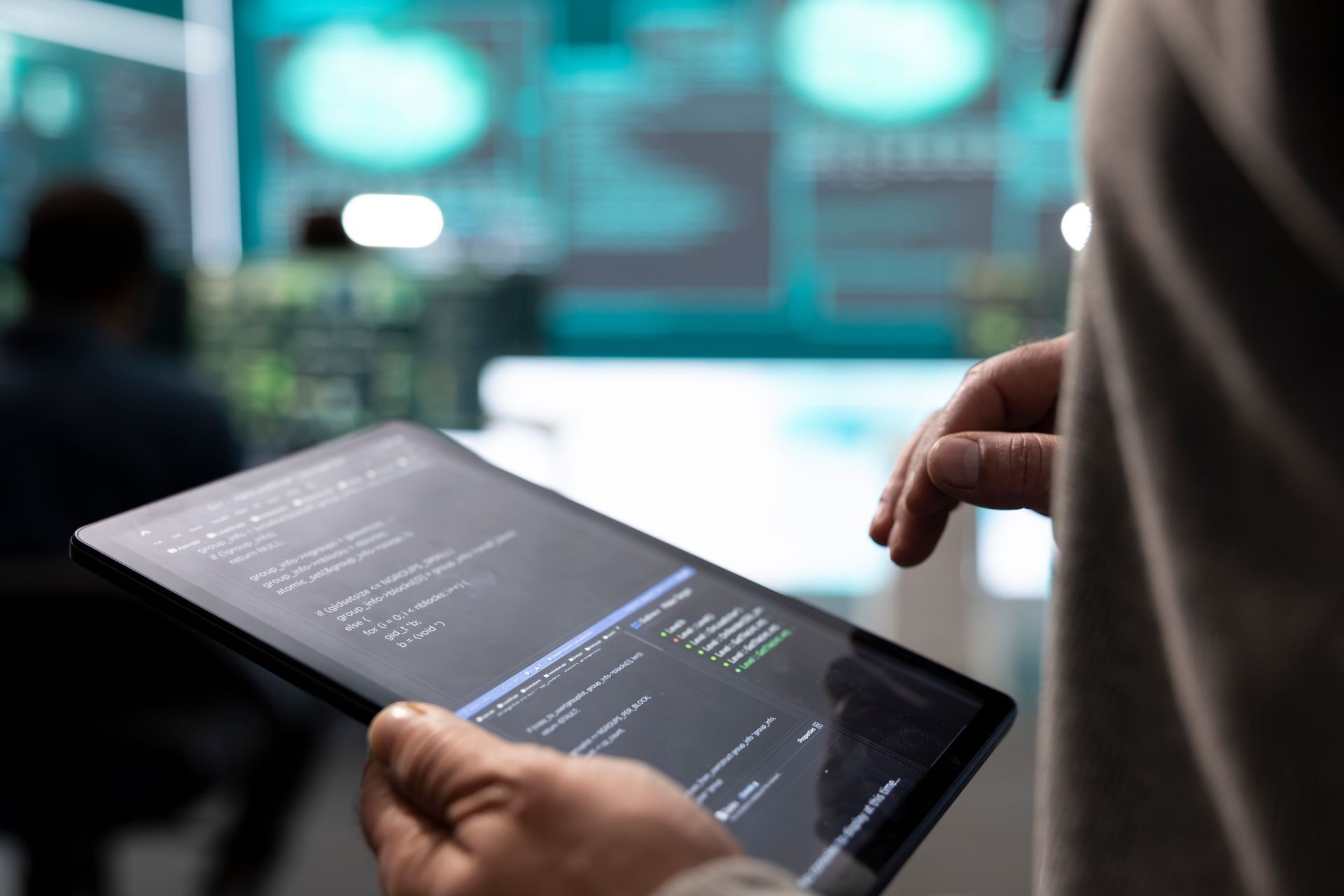 Persona sosteniendo una tableta con código, en una sala de control llena de tecnología.