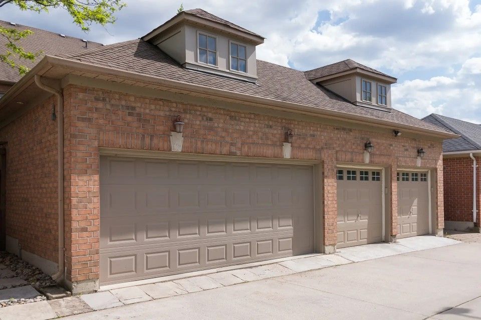 Garage en briques à trois portes, bardage beige et deux lucarnes sous un ciel nuageux.