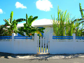 Maison blanche avec clôture et portail bleus, hautes plantes vertes et ciel d'un bleu éclatant.