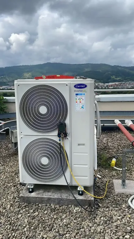 Unidad exterior de aire acondicionado con dos ventiladores sobre un tejado de grava, con montañas y un cielo nublado al fondo