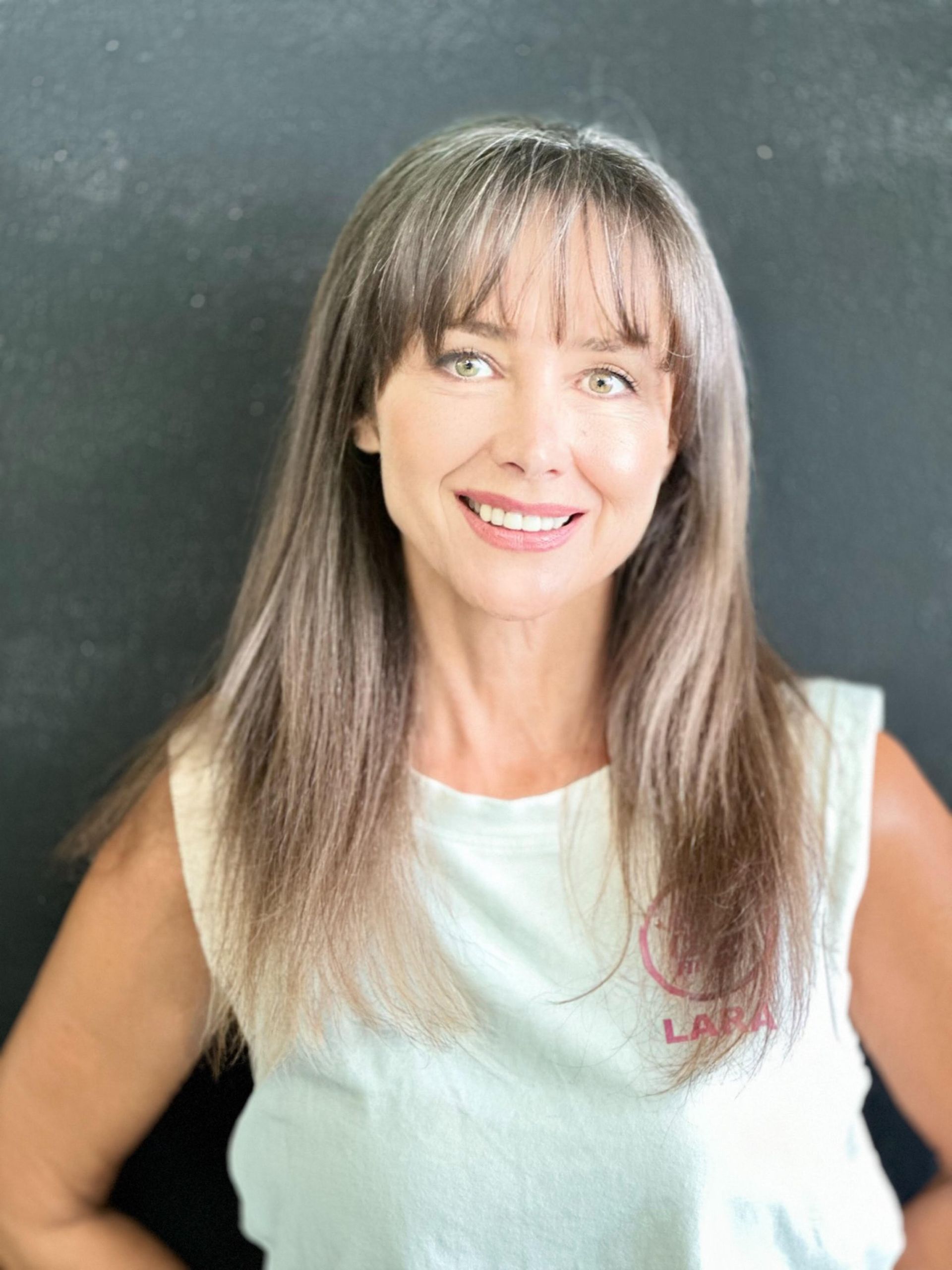 Una mujer con cabello largo y flequillo lleva una camiseta blanca sin mangas y sonríe.