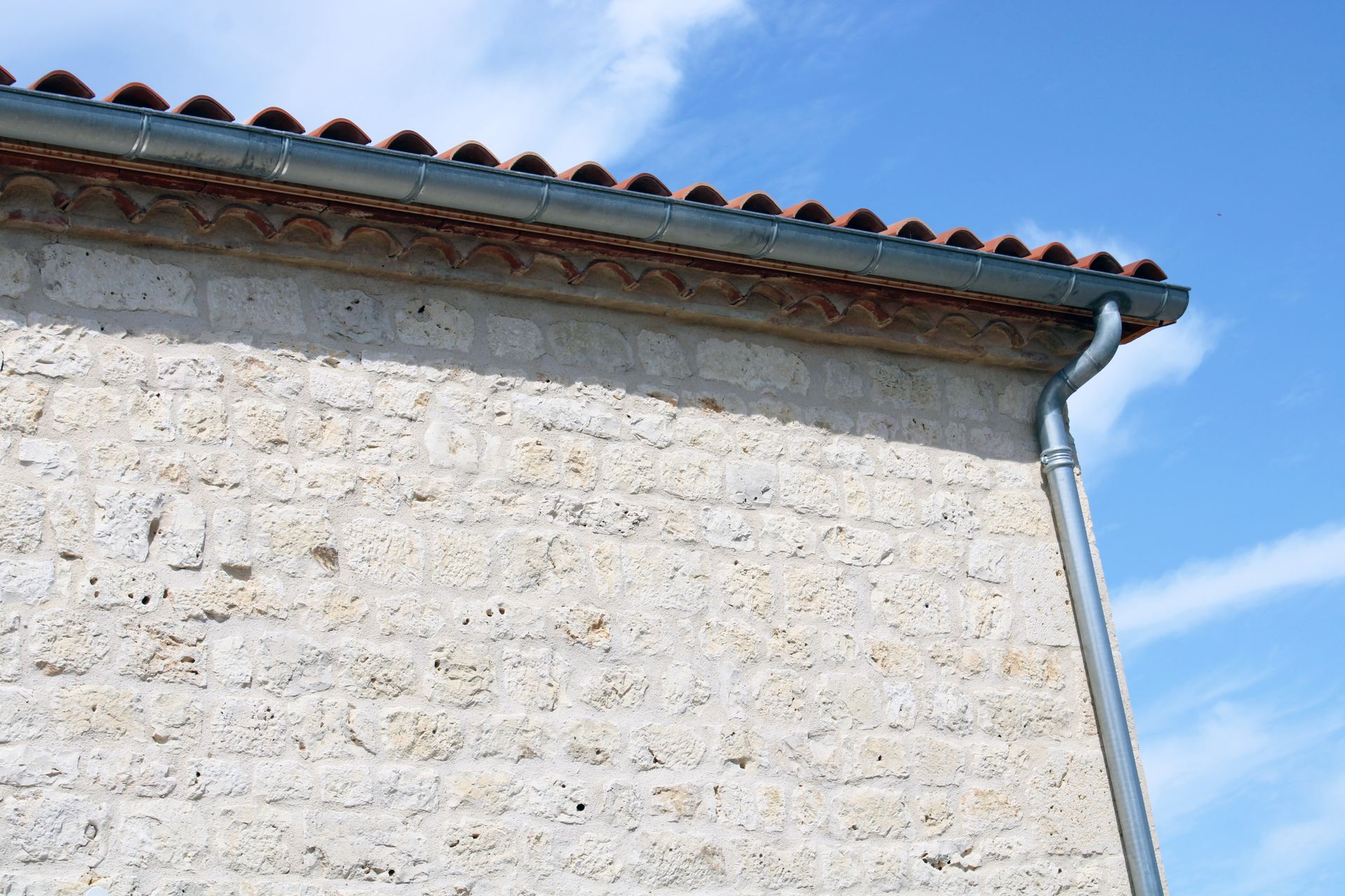 Coin d'un bâtiment en pierre avec une gouttière métallique et un toit de tuiles.