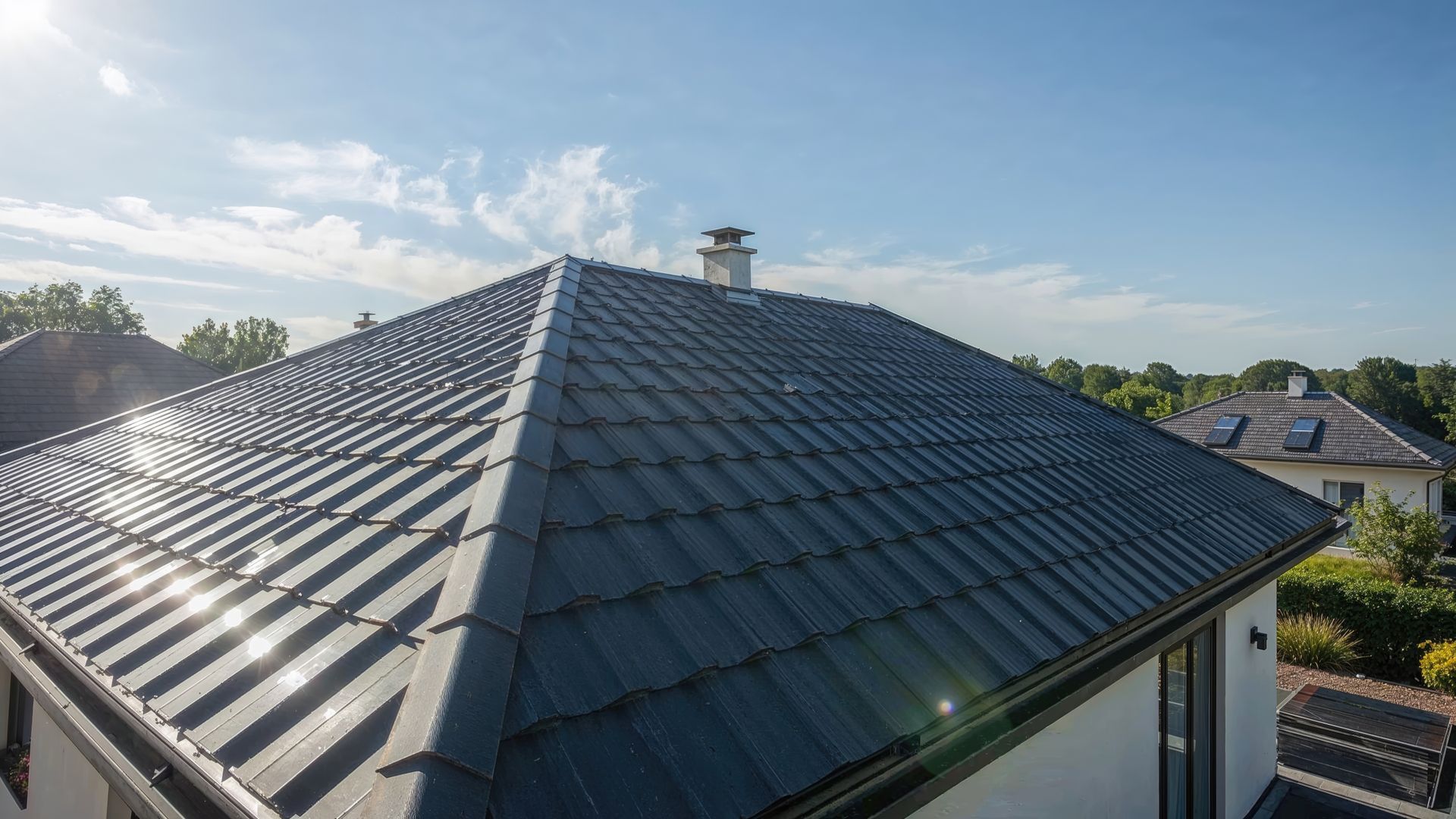 Toit de tuiles gris foncé sur une maison, cheminée et ciel bleu.