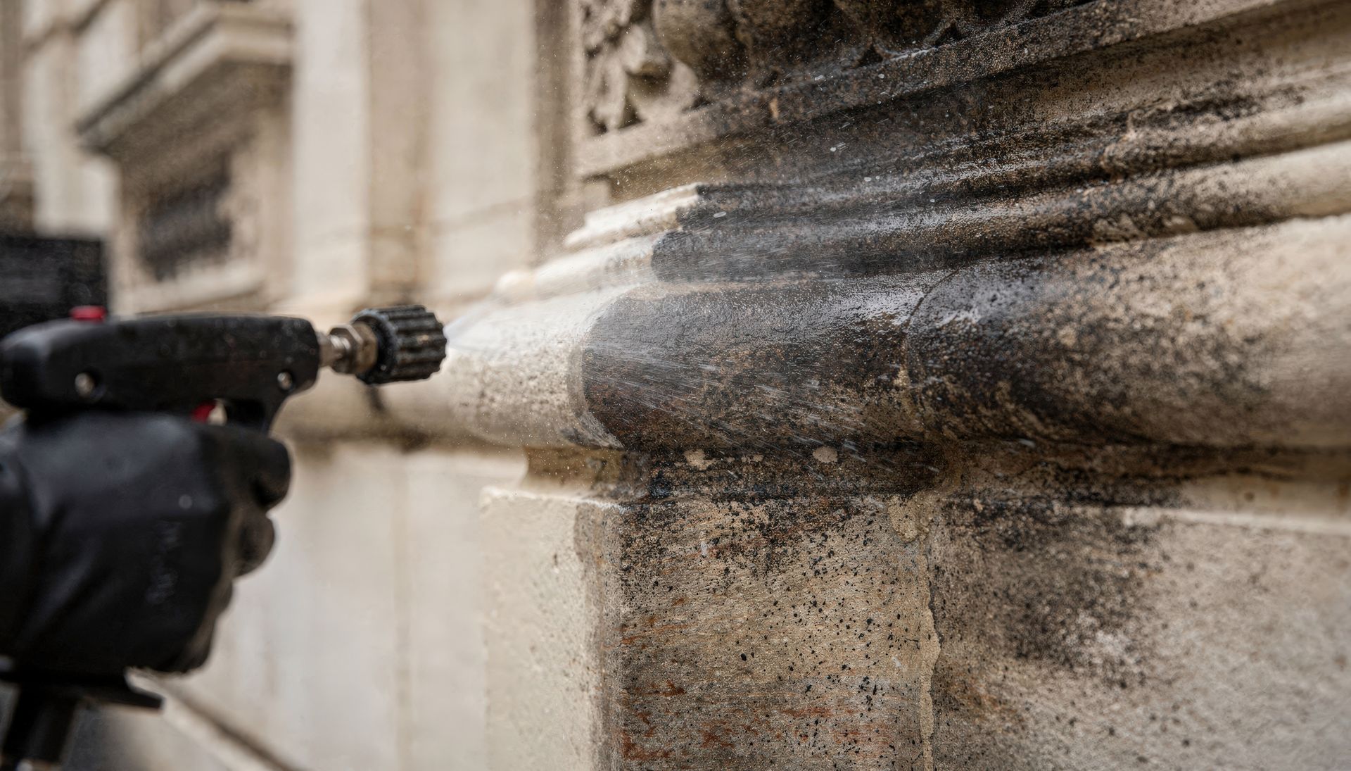 Une personne portant un gant noir nettoie à haute pression la façade en pierre d'un bâtiment.