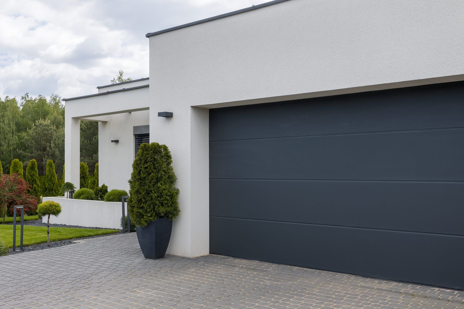 Porte de garage grise moderne sur une maison blanche, avec une allée gris foncé et une grande plante en pot.