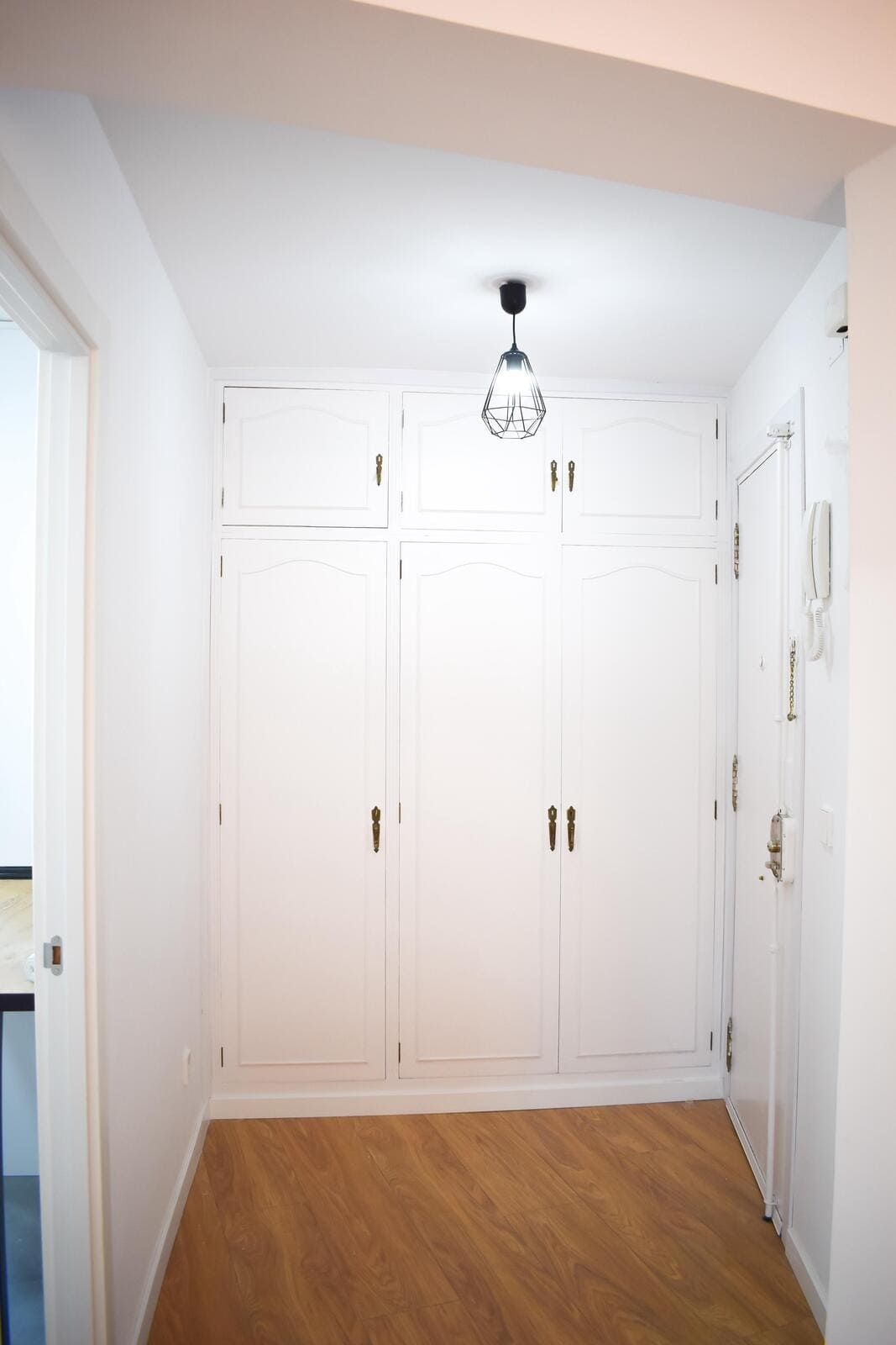 Pasillo blanco con suelo de madera y armarios empotrados blancos. Una lámpara negra cuelga del techo.