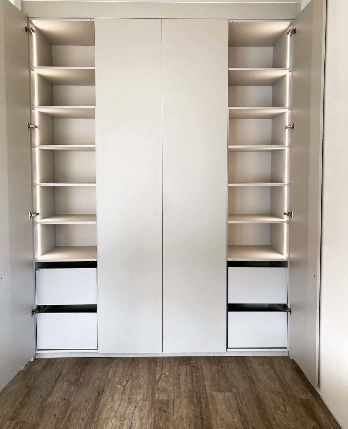 Armario empotrado blanco con puertas abiertas, estantes, cajones e iluminación interior, sobre un suelo de imitación madera clara.