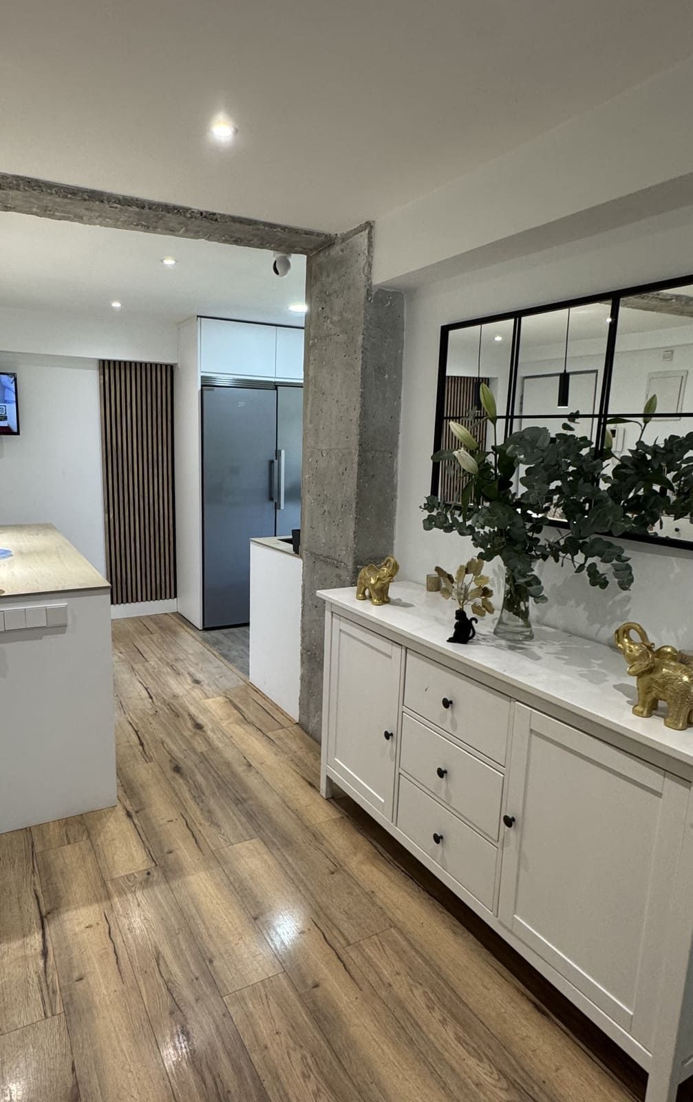 Cocina blanca con pisos de madera, gabinetes, espejo y refrigerador de acero inoxidable.