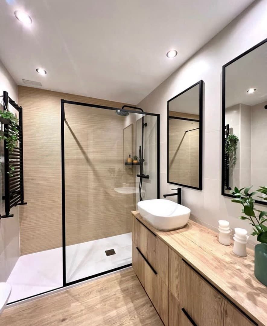 Baño moderno con ducha de cristal, tocador de madera y espejos con marco negro.