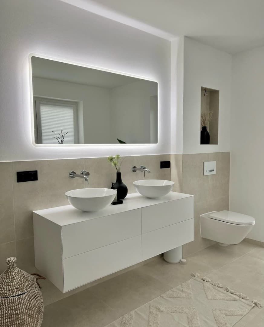 Baño blanco moderno con tocador flotante, lavabos sobre encimera, espejo retroiluminado y estante empotrado.