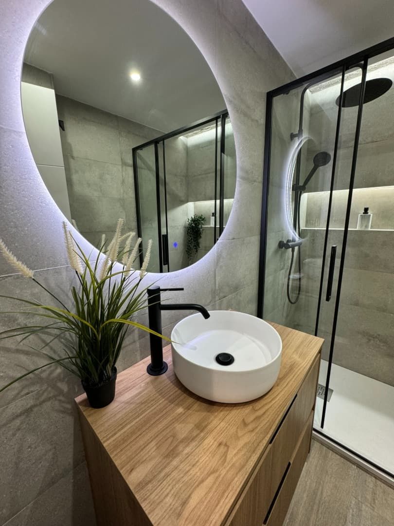 Baño moderno con tocador de madera, lavabo redondo, grifería negra, espejo iluminado y ducha de cristal.