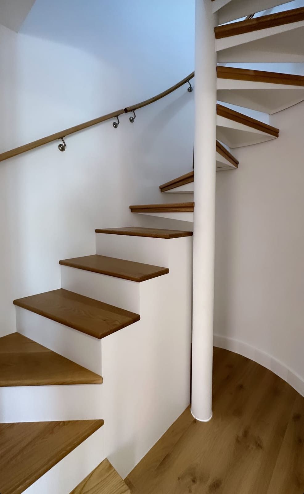 Escalera de caracol con escalones de madera y pasamanos contra paredes blancas.