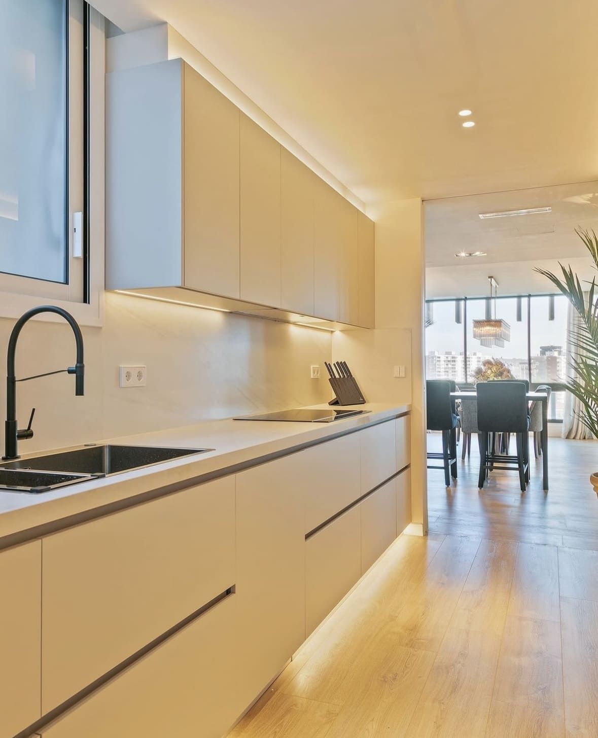 Cocina moderna con gabinetes claros, grifería negra e iluminación bajo los gabinetes. Comedor al fondo.