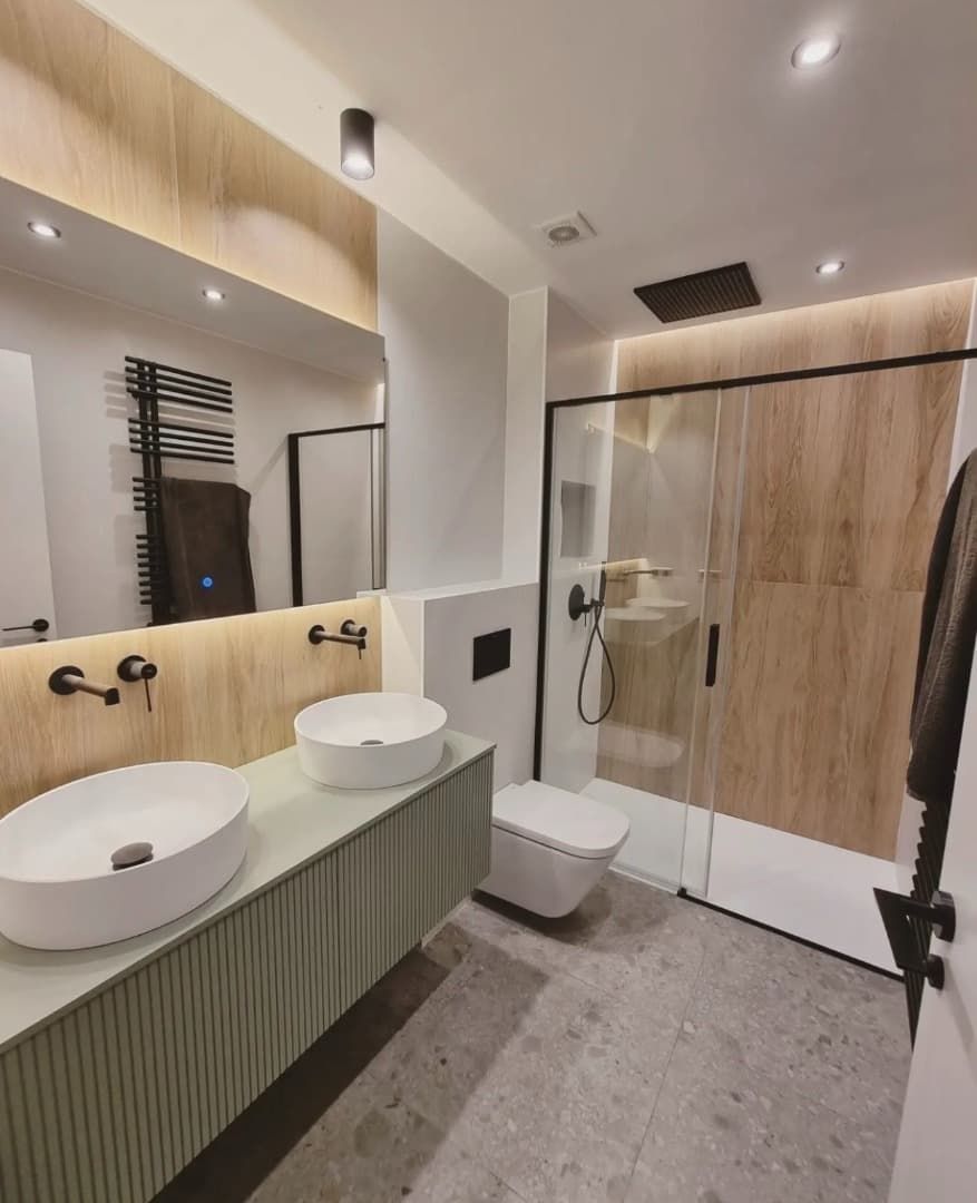 Baño moderno con lavabo doble, ducha e inodoro. Revestimiento de madera clara, suelo de baldosas grises y grifería negra.