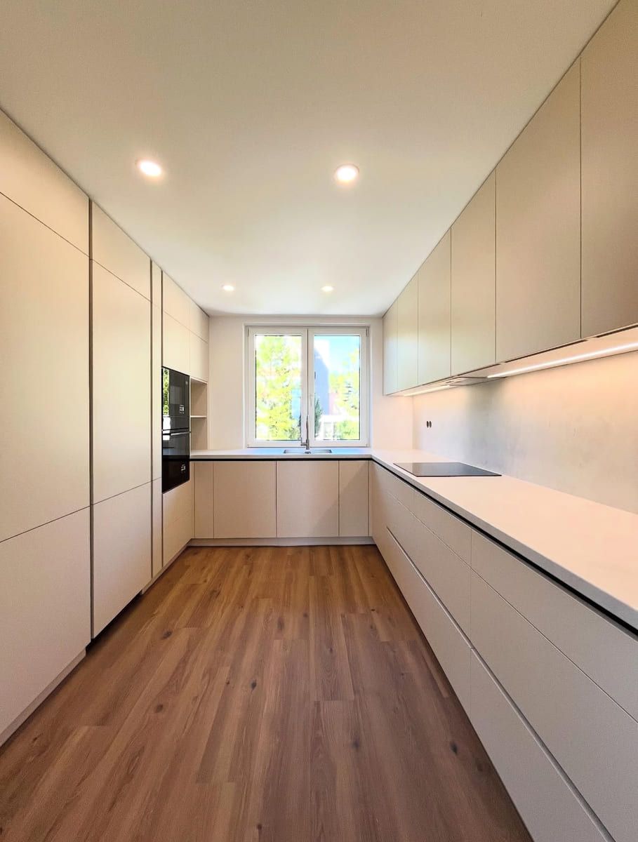 Cocina moderna en tonos neutros con piso de madera clara, gabinetes y una ventana.