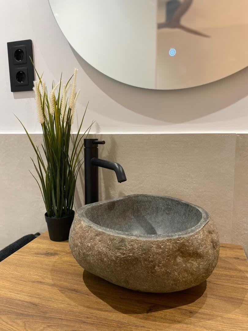 Baño con lavabo de piedra, grifería negra, encimera de madera, espejo y planta.