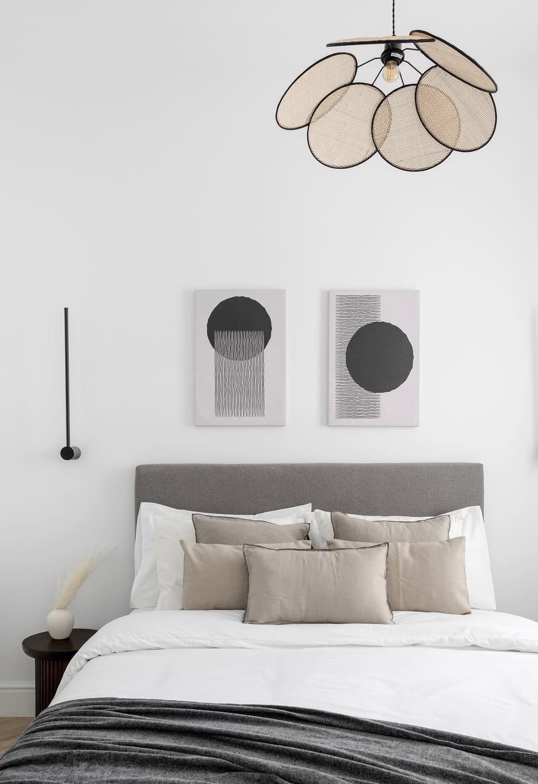 Dormitorio con cabecera gris, ropa de cama blanca y almohadas beige, con arte abstracto y una lámpara tejida.