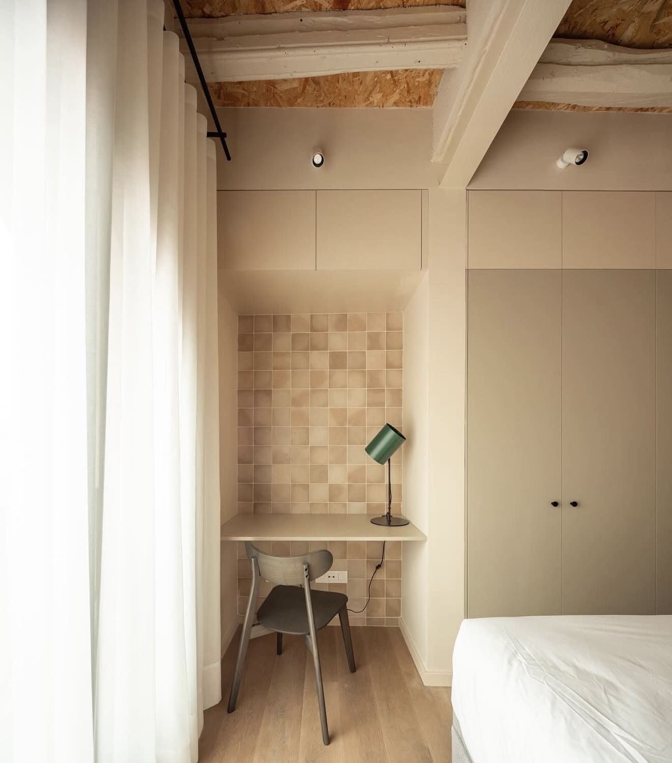 Dormitorio con escritorio empotrado, salpicadero de azulejos color canela, lámpara verde, silla gris y suelo de madera clara. Cortinas blancas y armario alto.