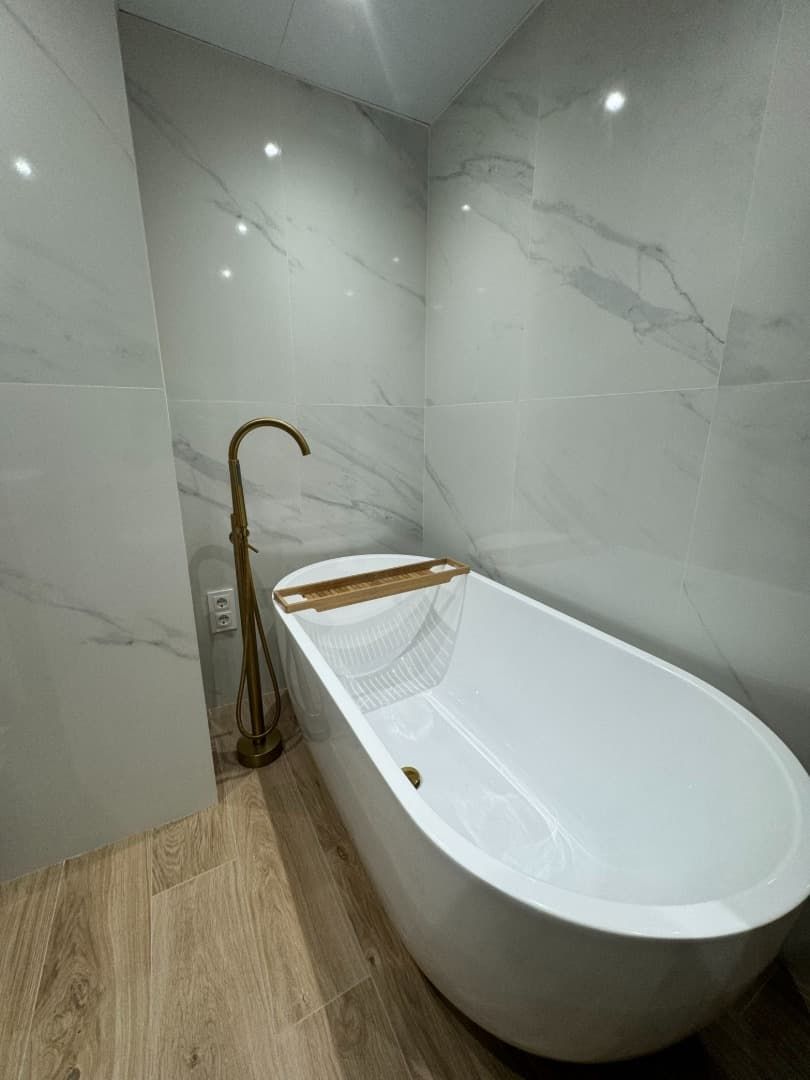 Una bañera ovalada blanca con grifo dorado en un baño con paredes estilo mármol y suelos imitación madera.
