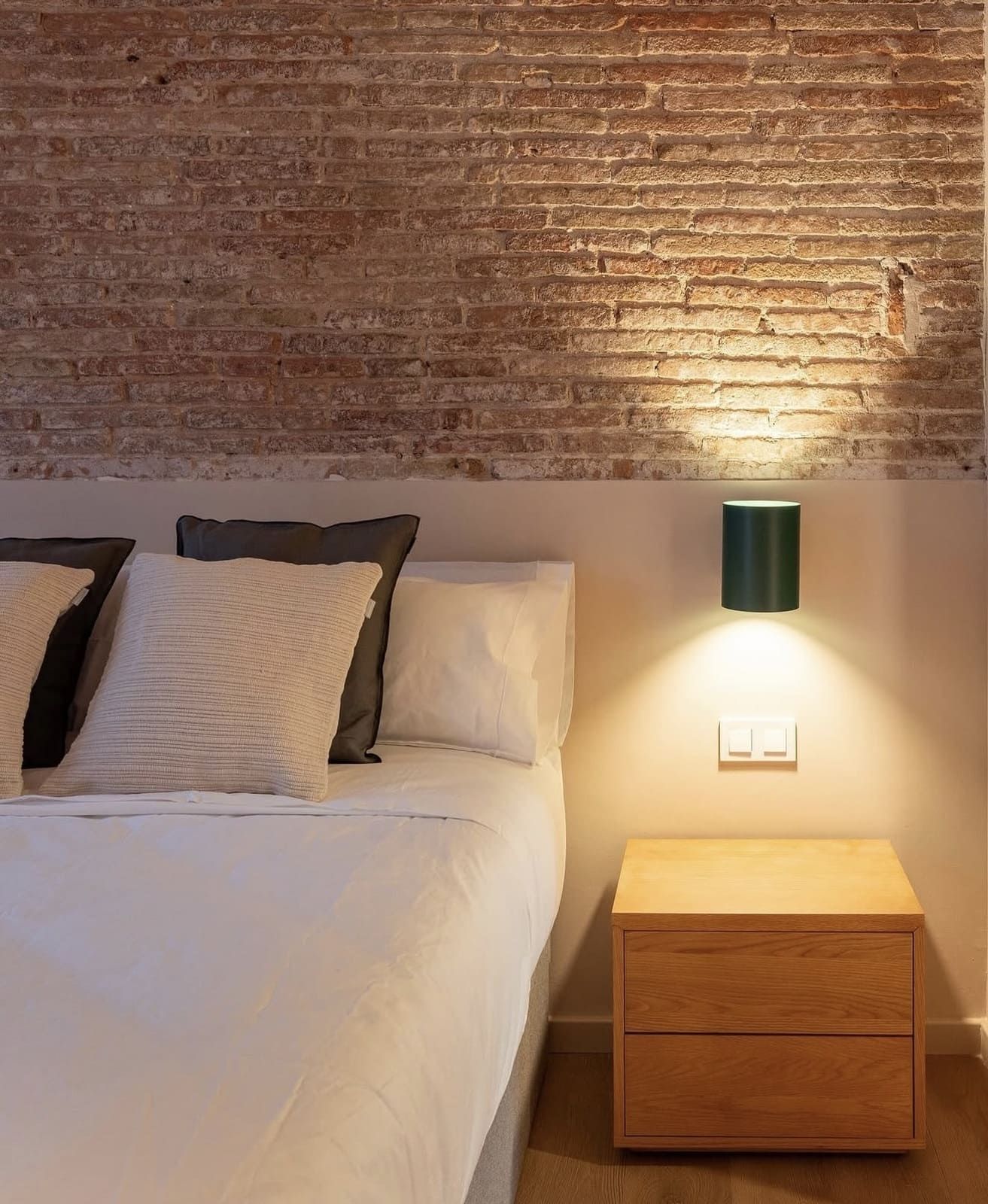 Dormitorio con pared de ladrillo y mesita de noche de madera, iluminada por un aplique. Ropa de cama y almohadas blancas sobre la cama.