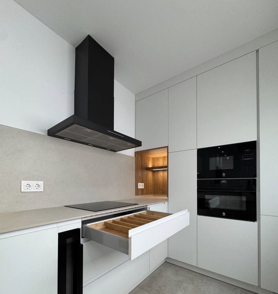 Cocina moderna con gabinetes blancos, campana negra, electrodomésticos empotrados y cajón abierto con utensilios.