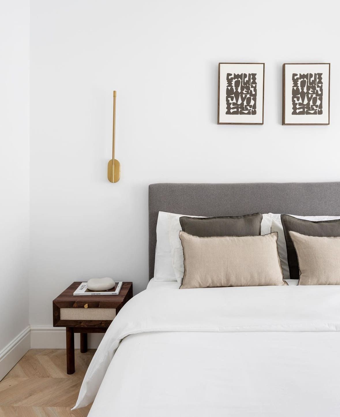 Dormitorio con cabecero gris, ropa de cama blanca, mesita de noche de madera y láminas artísticas.