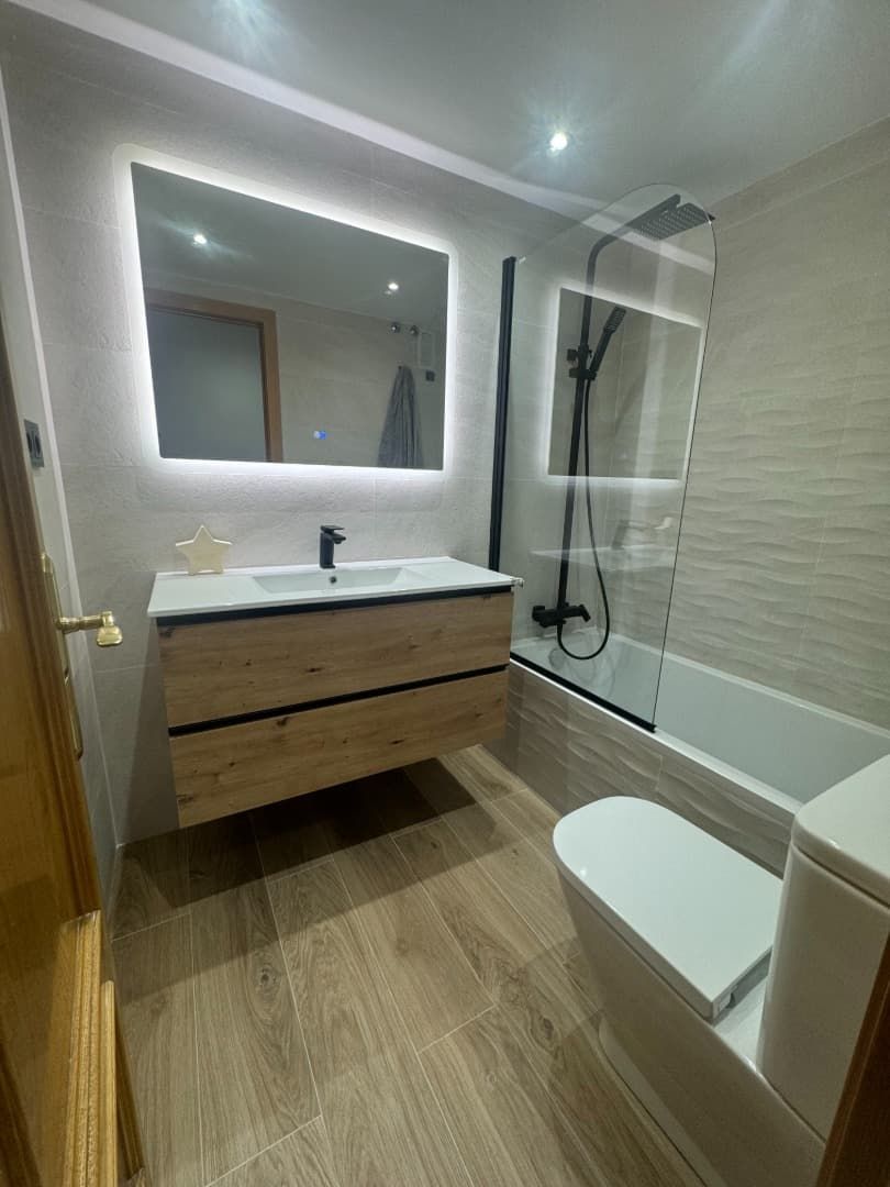 Baño moderno con tocador imitación madera, espejo retroiluminado, mampara de cristal y suelo de baldosas.