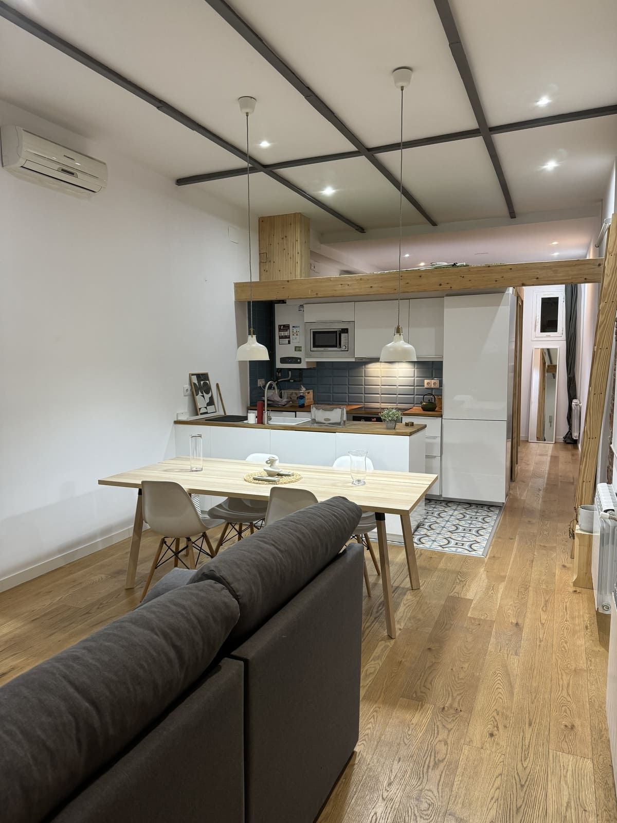 Acogedora cocina moderna con altillo. Armarios blancos, mesa de madera clara y sofá gris. Luz natural y suelos de madera.