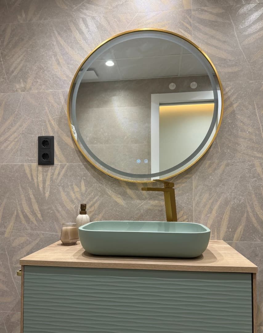 Tocador de baño con espejo redondo, grifo dorado y lavabo rectangular verde, sobre un fondo de azulejos florales.