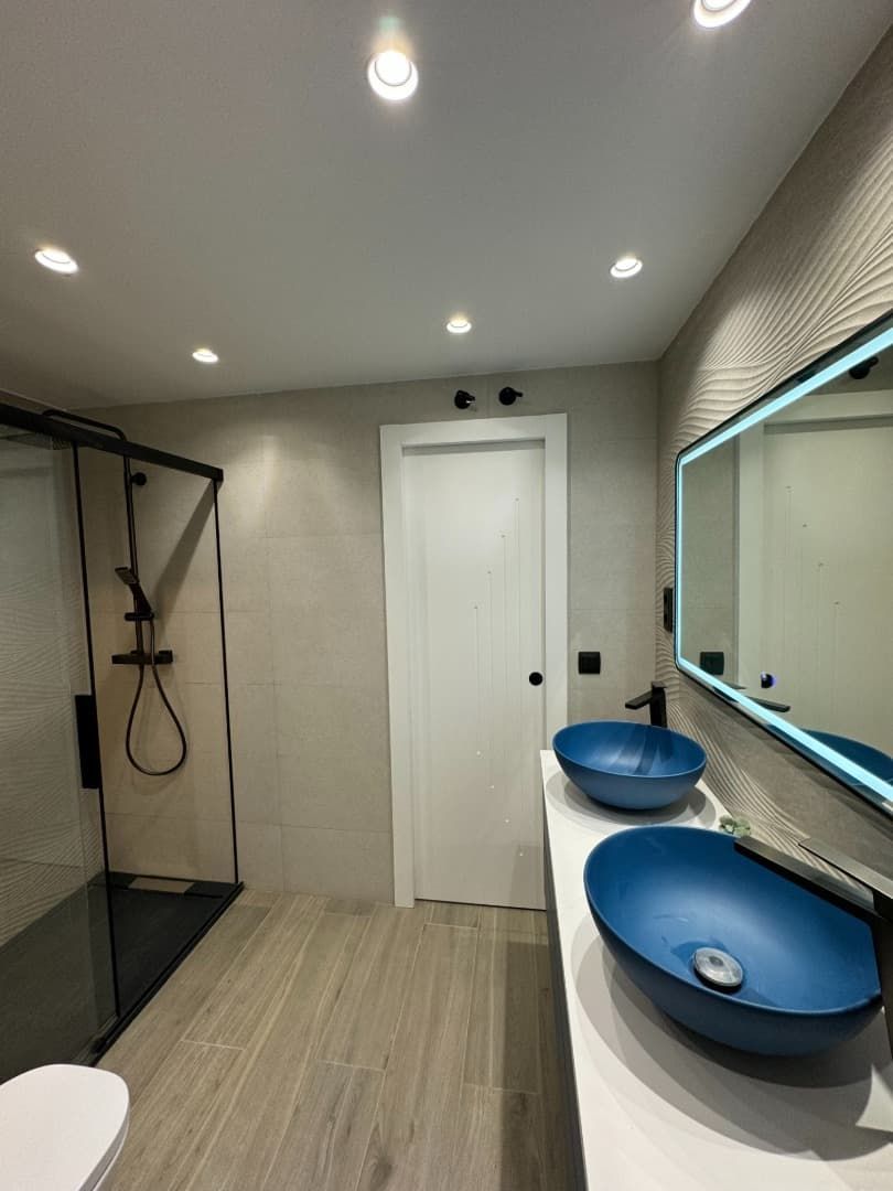 Baño moderno con lavabos de color azul, espejo retroiluminado, ducha y puerta blanca.