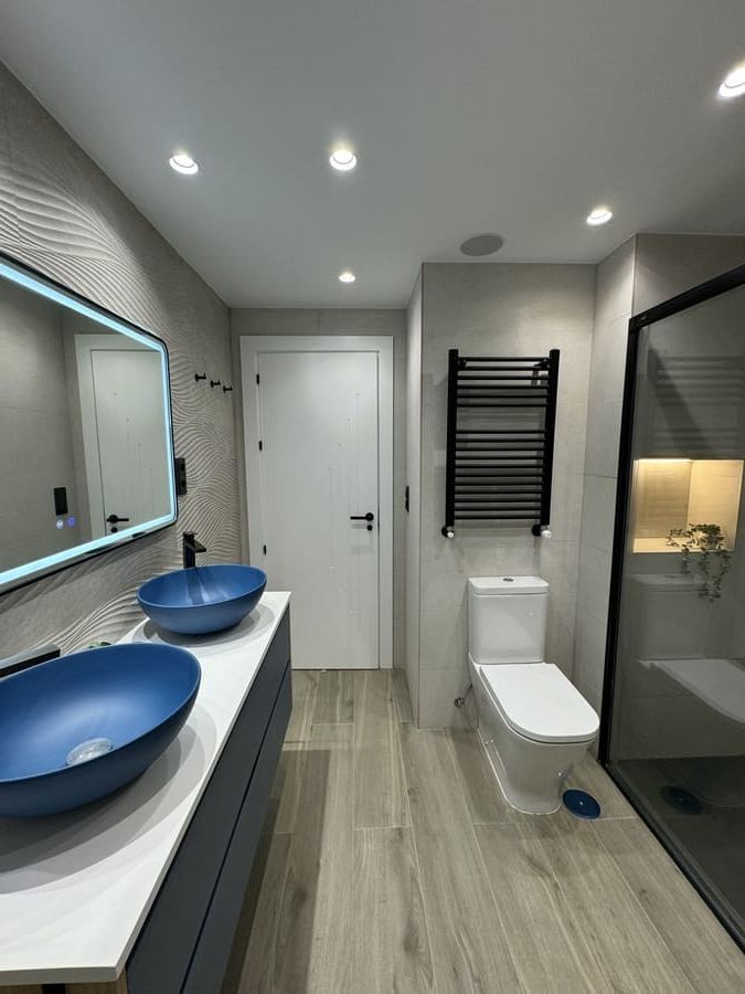 Baño moderno con tocador de madera, espejo redondo, ducha de cristal y paredes de azulejos grises.