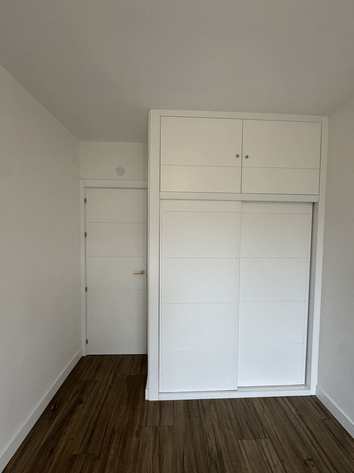 Armario empotrado blanco con puertas corredizas, junto a una puerta blanca cerrada en una habitación con suelo de madera.