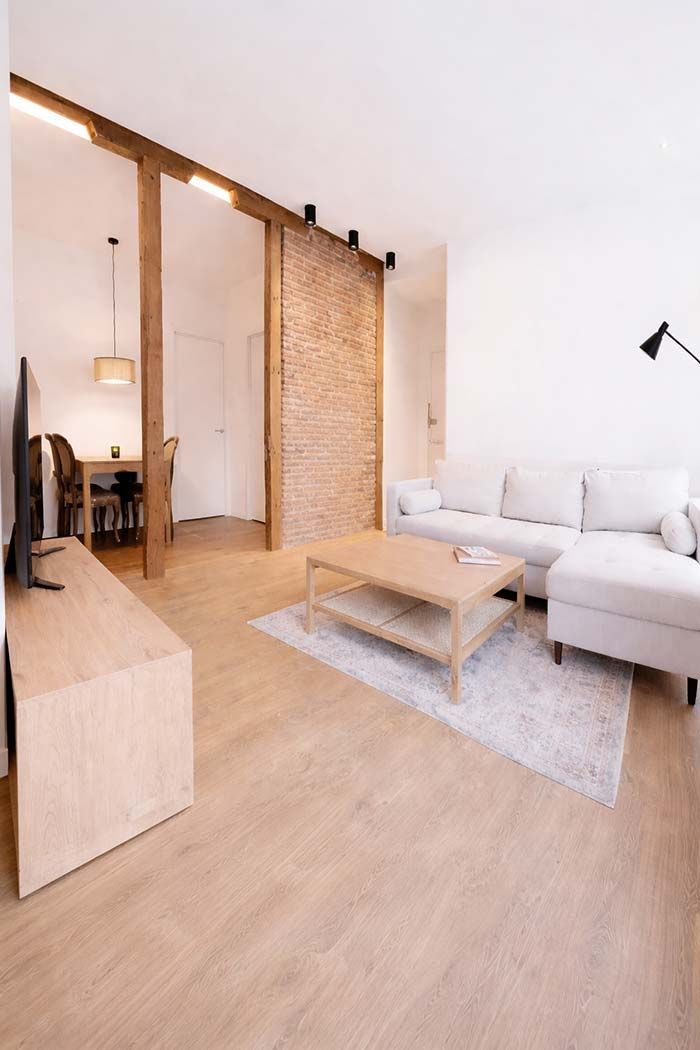 Sala de estar moderna con cocina. Sofá gris, suelo de madera, isla con taburetes e iluminación cenital.