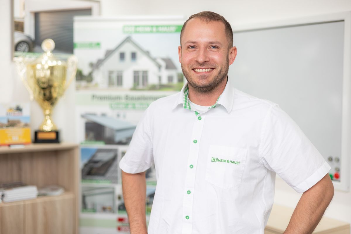 Ein Mann in einem weißen Hemd steht vor einem Haus und einer Trophäe.