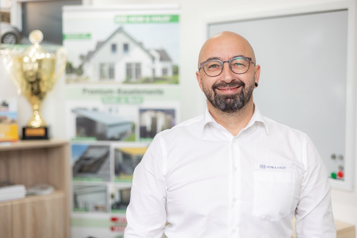 Ein Mann mit Bart und Brille trägt ein weißes Hemd und lächelt.