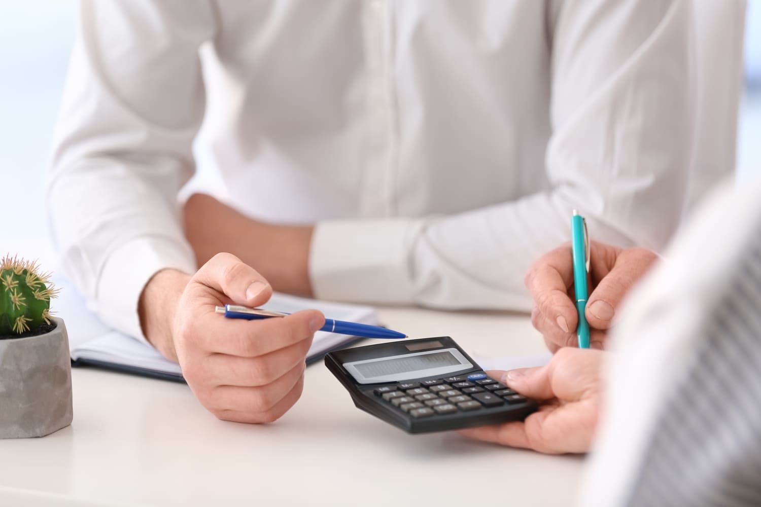 Dos personas sentadas en un escritorio usando una calculadora y escribiendo. Cerca hay un pequeño cactus en una maceta.