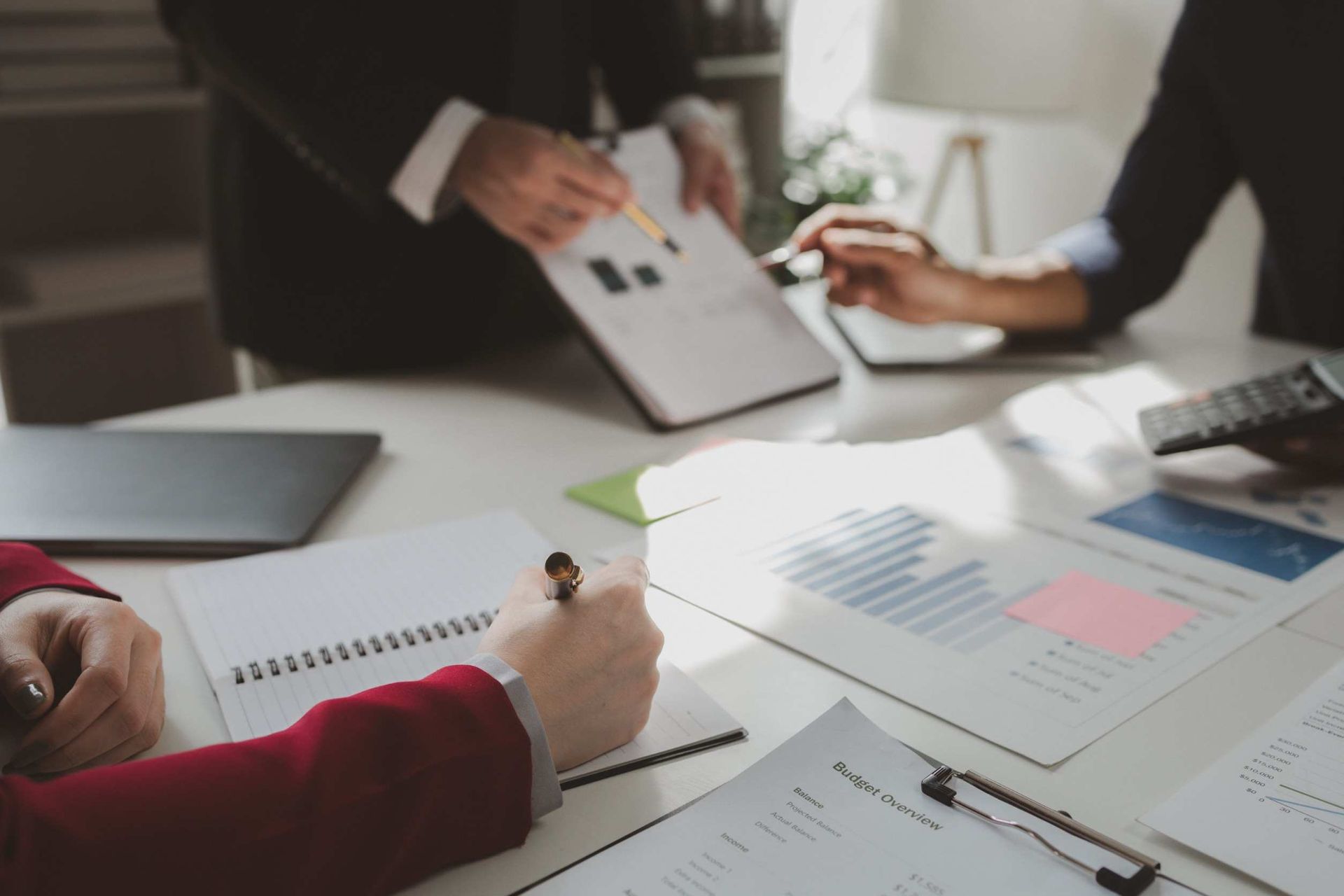 Empresarios en una mesa revisando informes financieros. Uno escribe en un cuaderno, mientras otros señalan gráficos y tablas.