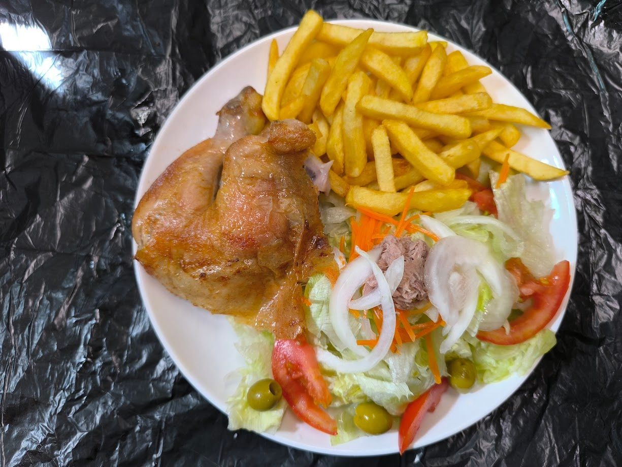 Plato con muslo de pollo asado, patatas fritas y ensalada que incluye tomates, aceitunas y cebollas.