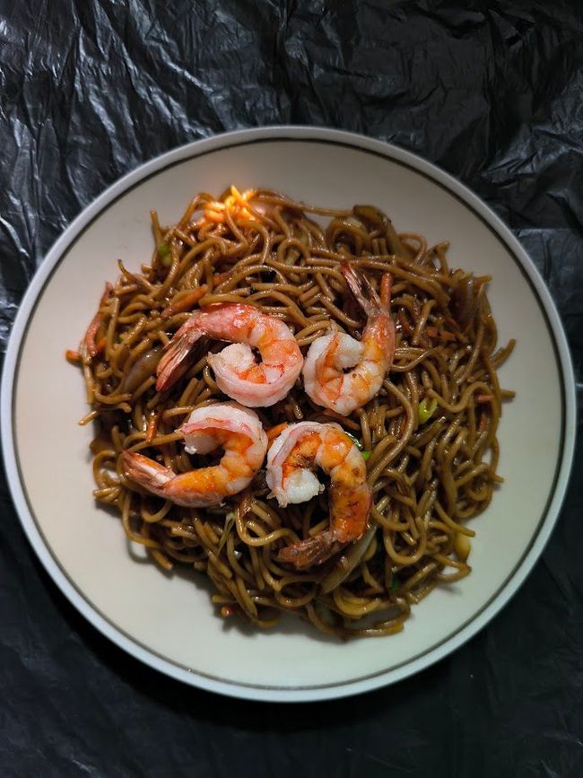 Plato de fideos con cuatro camarones cocidos encima. Los fideos son oscuros y están espolvoreados con semillas de sésamo. El plato está sobre una superficie negra.