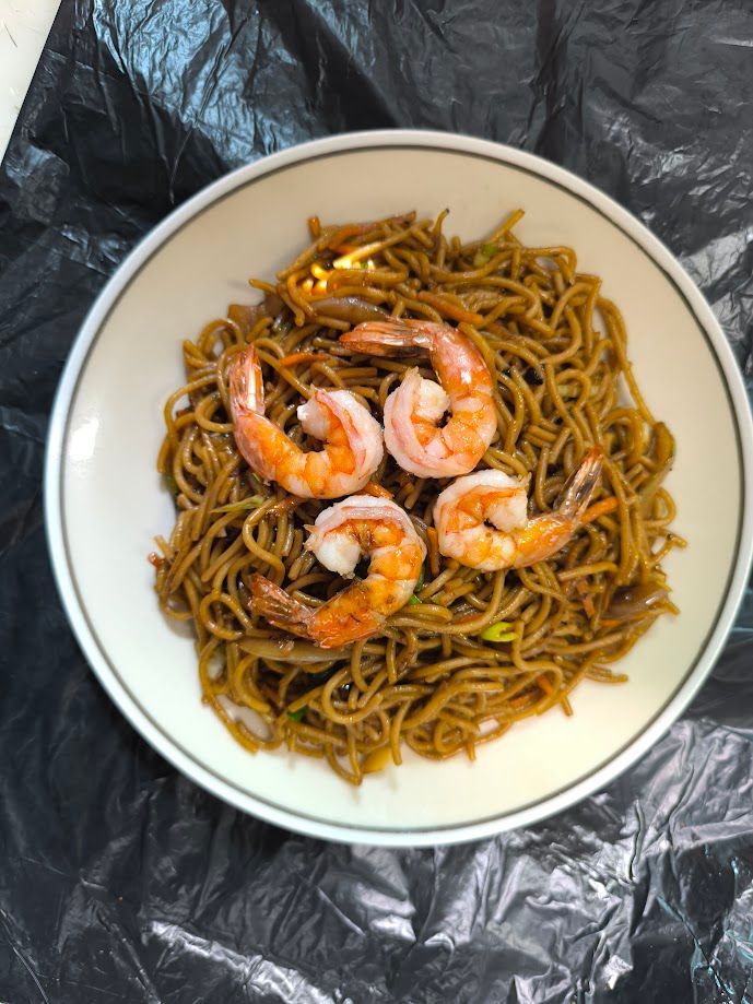 Un recipiente con fideos con cuatro camarones cocidos dispuestos encima, contra una superficie negra.