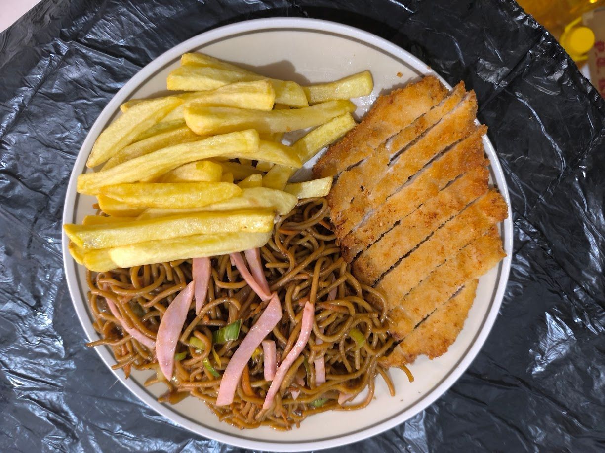 Plato de comida que incluye patatas fritas, fideos fritos con tiras rosadas y una chuleta rebozada y cortada en rodajas.