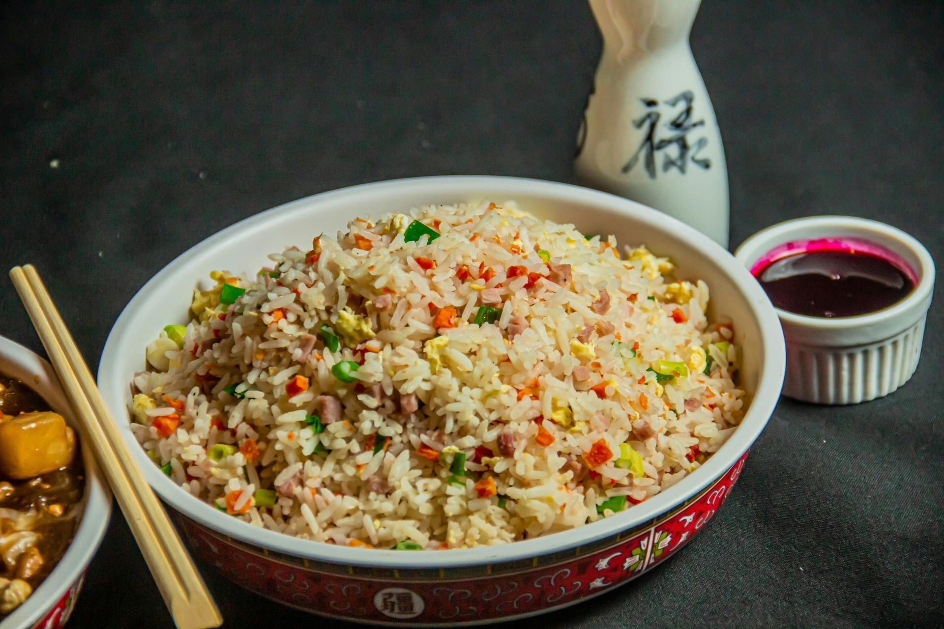 Tazón de arroz frito con zanahorias, guisantes y huevo, con palillos, salsa y una botella de sake sobre un fondo oscuro.